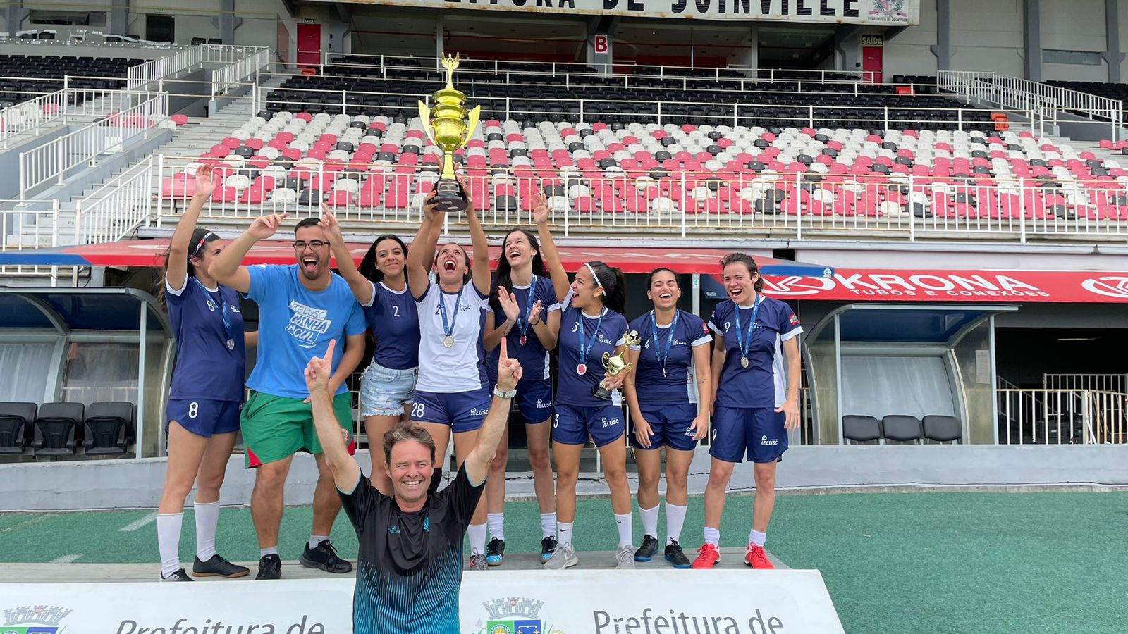 Uniformizados, alunas e alunos da Faculdade Ielusc erguem um troféu dourado, simbolizando a conquista do Jeville 2022.