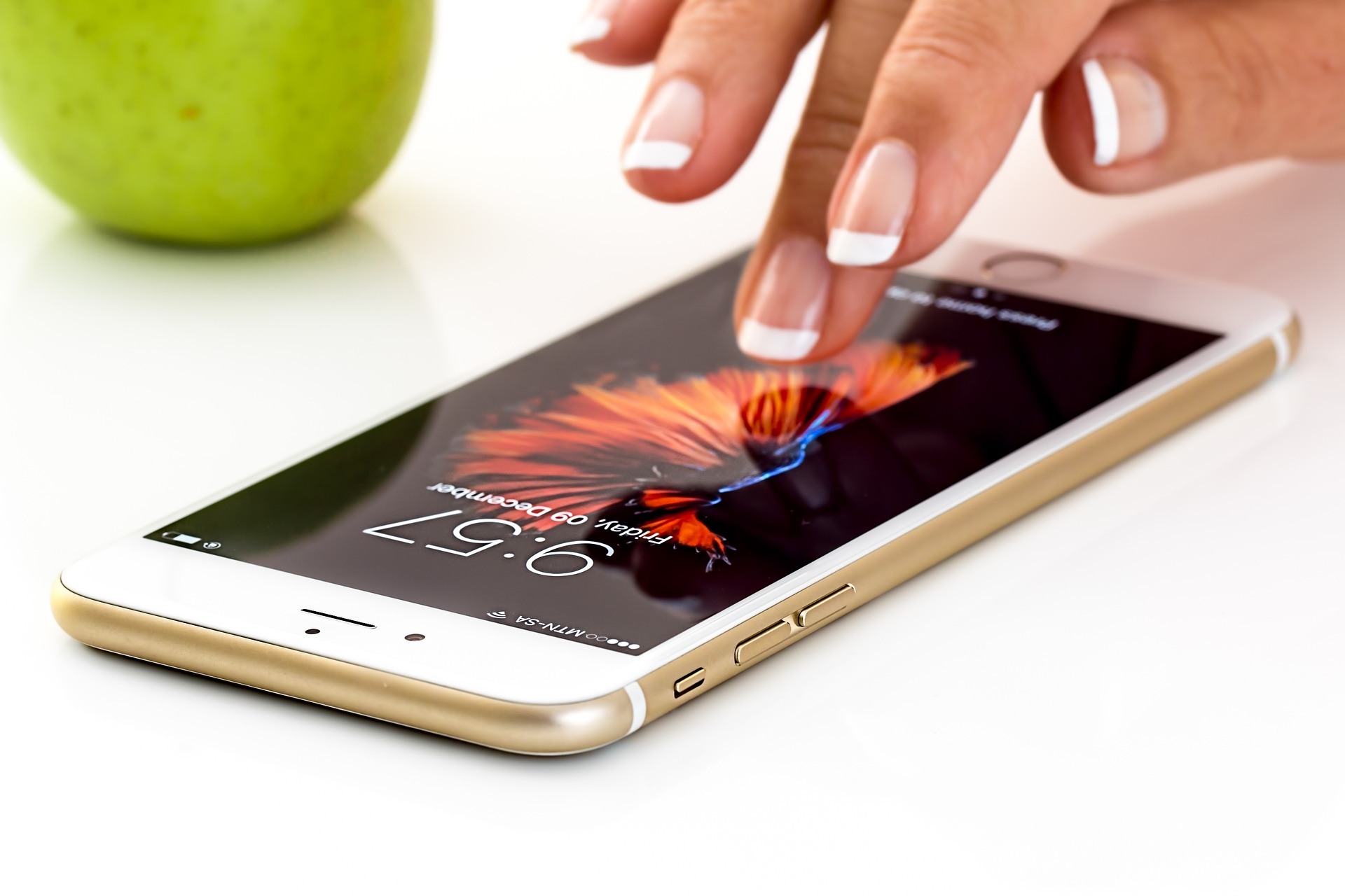 Mão, com unhas pintadas em "francesinha", mexendo em um celular com uma maçã verde ao fundo.