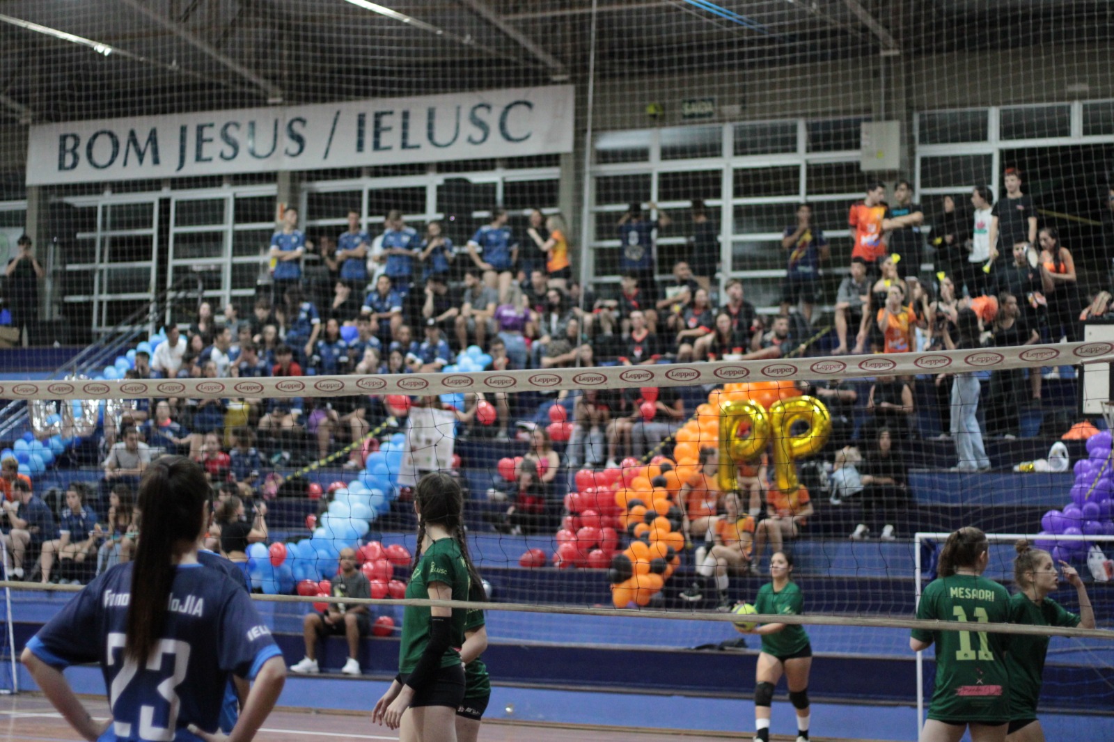 Atletas dos cursos de Nutrição, com camisas verdes, e Fonoaudiologia, com camisas azuis, em uma partida de vôlei no JIA. As equipes estão separadas por uma rede, e ao fundo estão as torcidas.