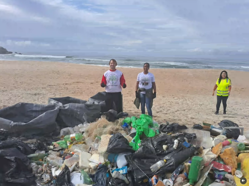 Montanha de resíduos plásticos, redes de pesca e sacos de lixo recolhidos durante ação de limpeza ocupa o primeiro plano da imagem. Ao fundo, três pessoas (as duas do centro são o casal de fundadores) estão em pé na faixa de areia de uma praia, com o mar e o céu nublado ao fundo.