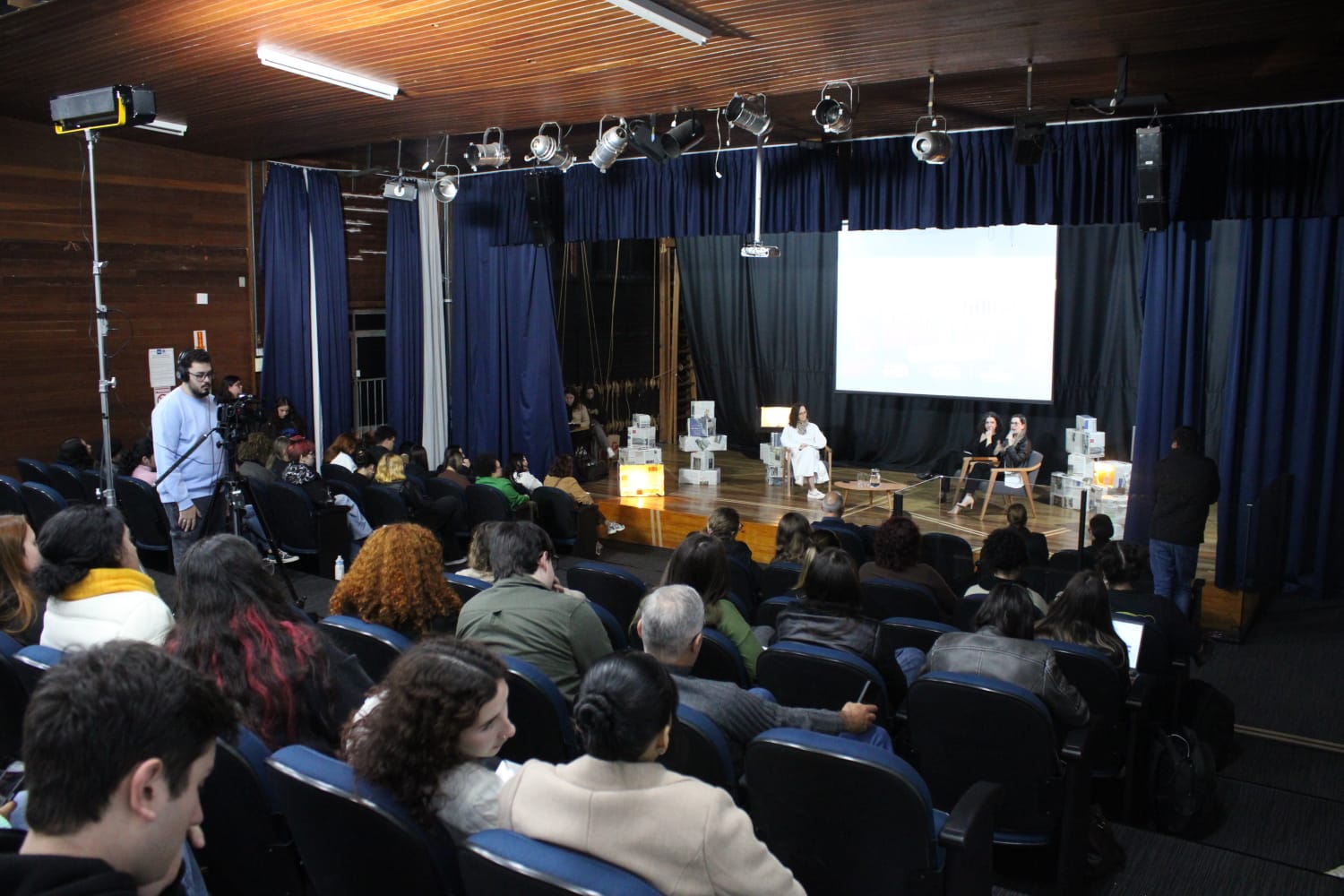 Fotografia colorida em plano geral do auditório do IELUSC mostra o público de traz para frente nas poltronas enquanto, no palco , ocorre o painel com as convidadas do evento.