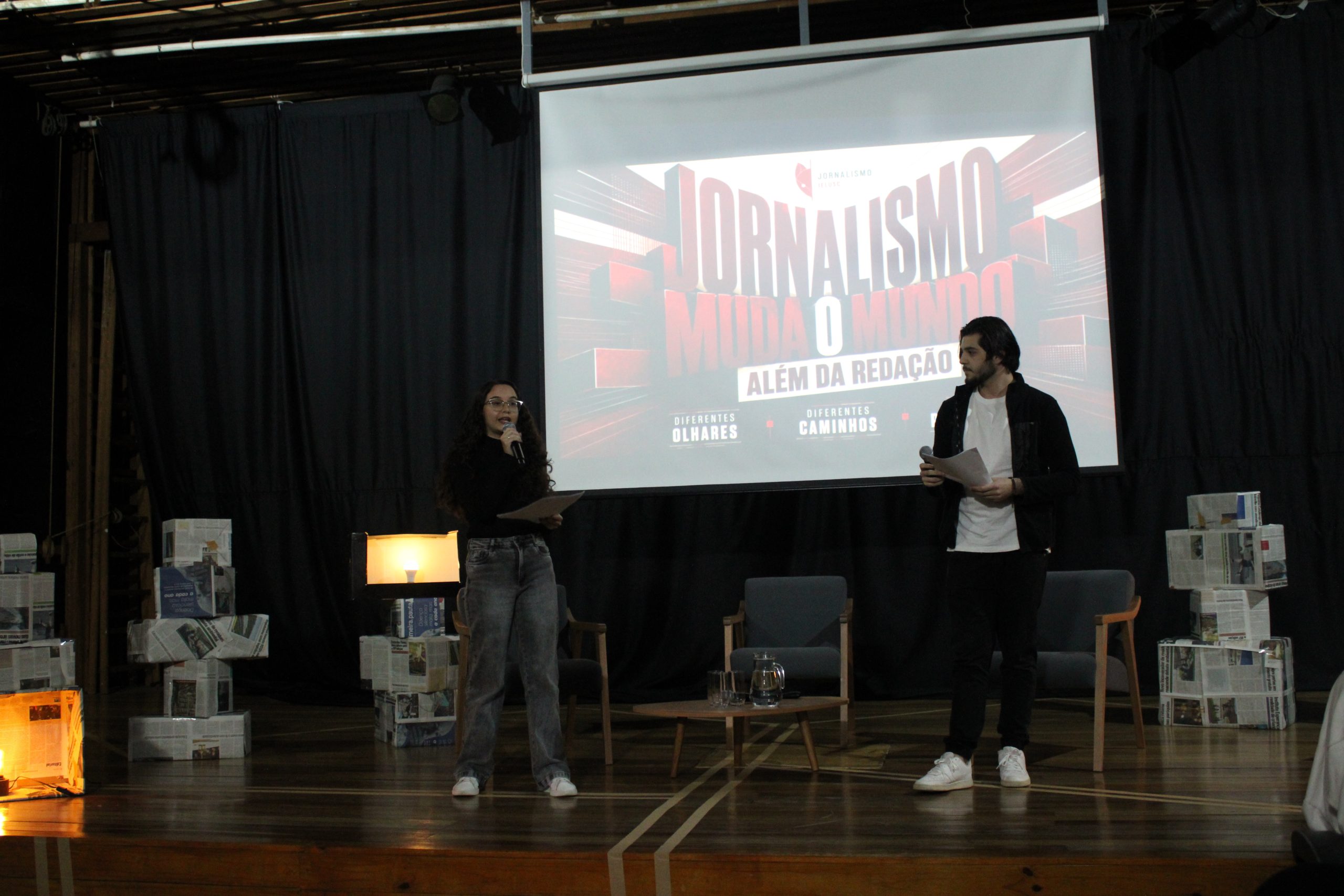 Fotografia colorida em plano aberto dos estudantes Giovanna Marques e Raulino Dalcim enquanto apresentam evento sobre jornalismo em palco decorado com luminárias e itens de jornais, diante de um telão com o tema 'Jornalismo muda o mundo: além da redação'