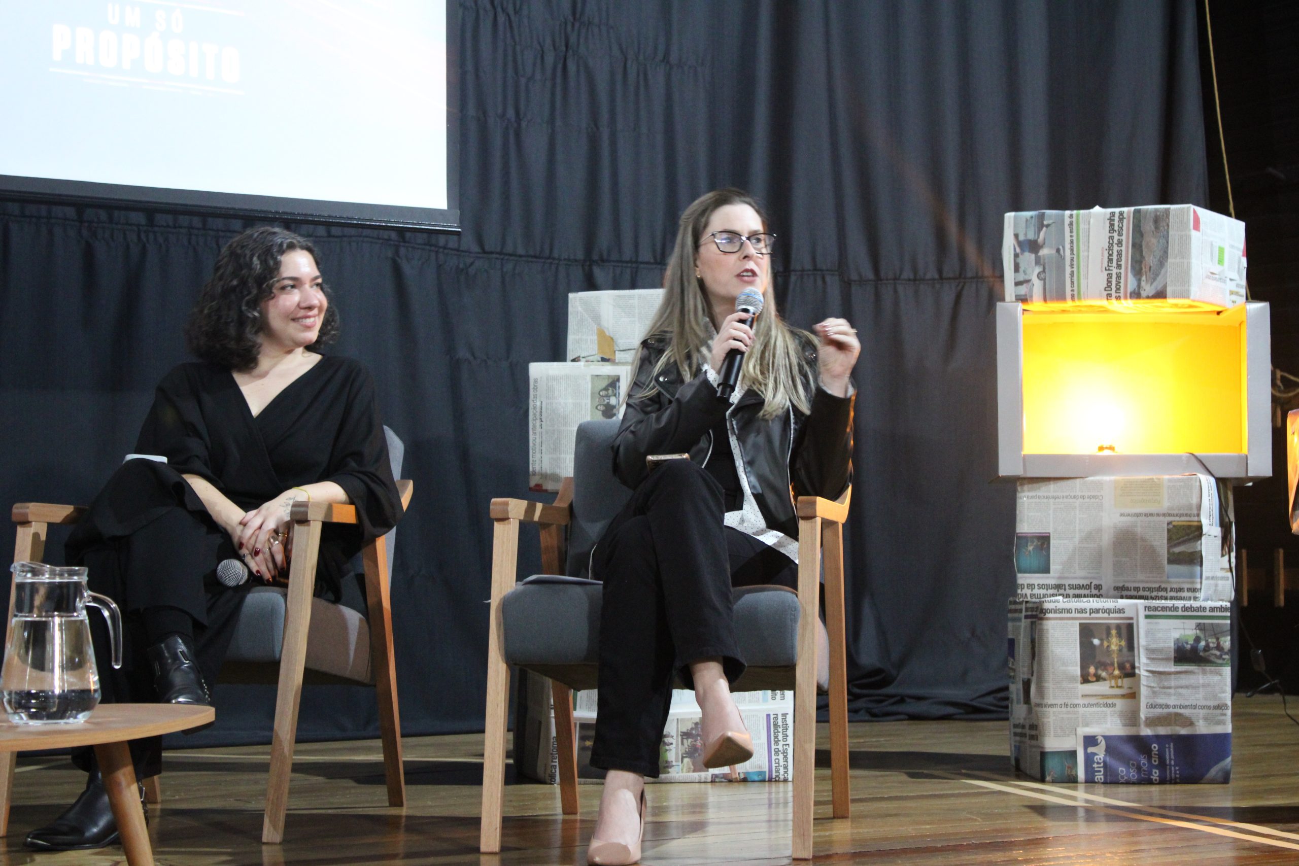 Fotografia em plano médio das convidadas Geórgia Santos e Graziéli Bottari sentadas em poltronas sobre um palco durante o evento. Geórgia Santos, à esquerda, veste roupa preta e observa a colega com um sorriso. Graziéli Bottari, ao centro, também de preto, segura um microfone enquanto fala ao público. Ao lado das convidadas há uma mesa com uma jarra de água e, à direita, uma luminária amarela apoiada sobre estruturas revestidas com jornais.