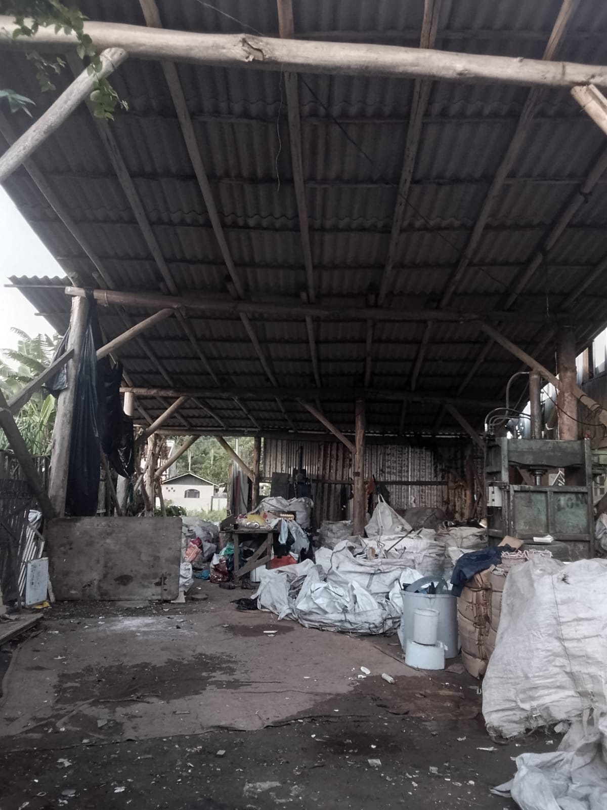 Galpão de triagem e armazenamento de materiais recicláveis com estrutura simples de madeira e cobertura de fibrocimento. Sacos de recicláveis, equipamentos de prensagem e materiais separados ocupam o interior do espaço.
