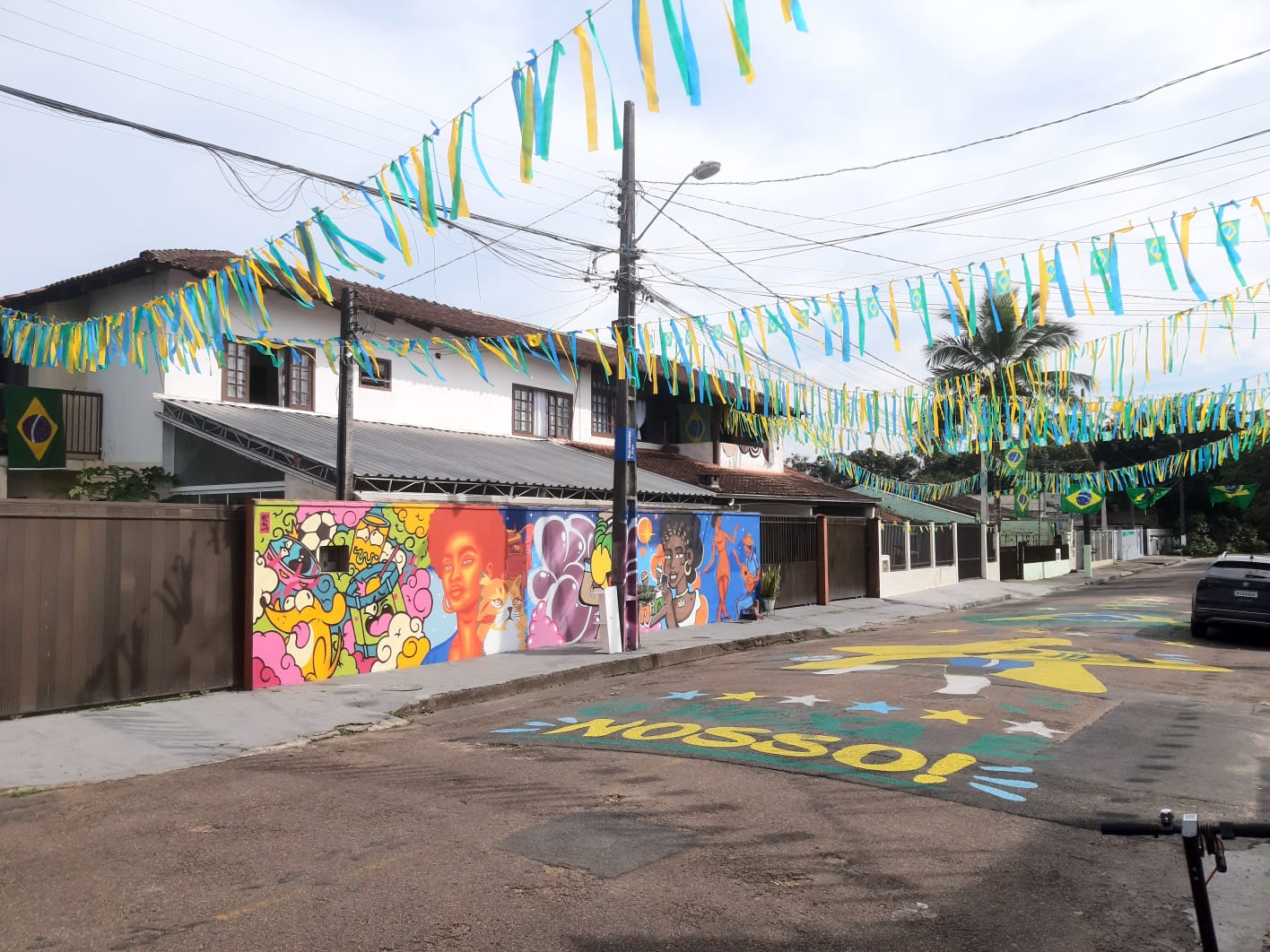 Vista lateral de rua residencial decorada para a Copa do Mundo, com bandeirolas suspensas, mural artístico em uma parede e pintura temática no asfalto 