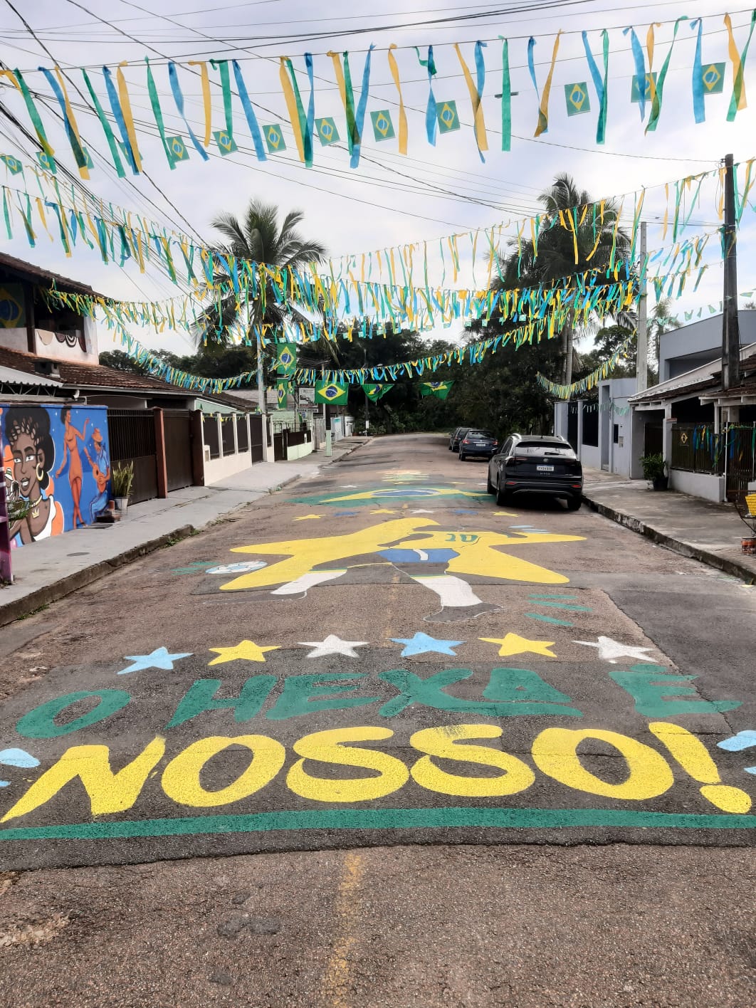 Rua decorada com bandeirolas verde-amarelas e pintura temática da Copa do Mundo no asfalto, com a palavra "Nosso" e elementos nas cores da bandeira brasileira