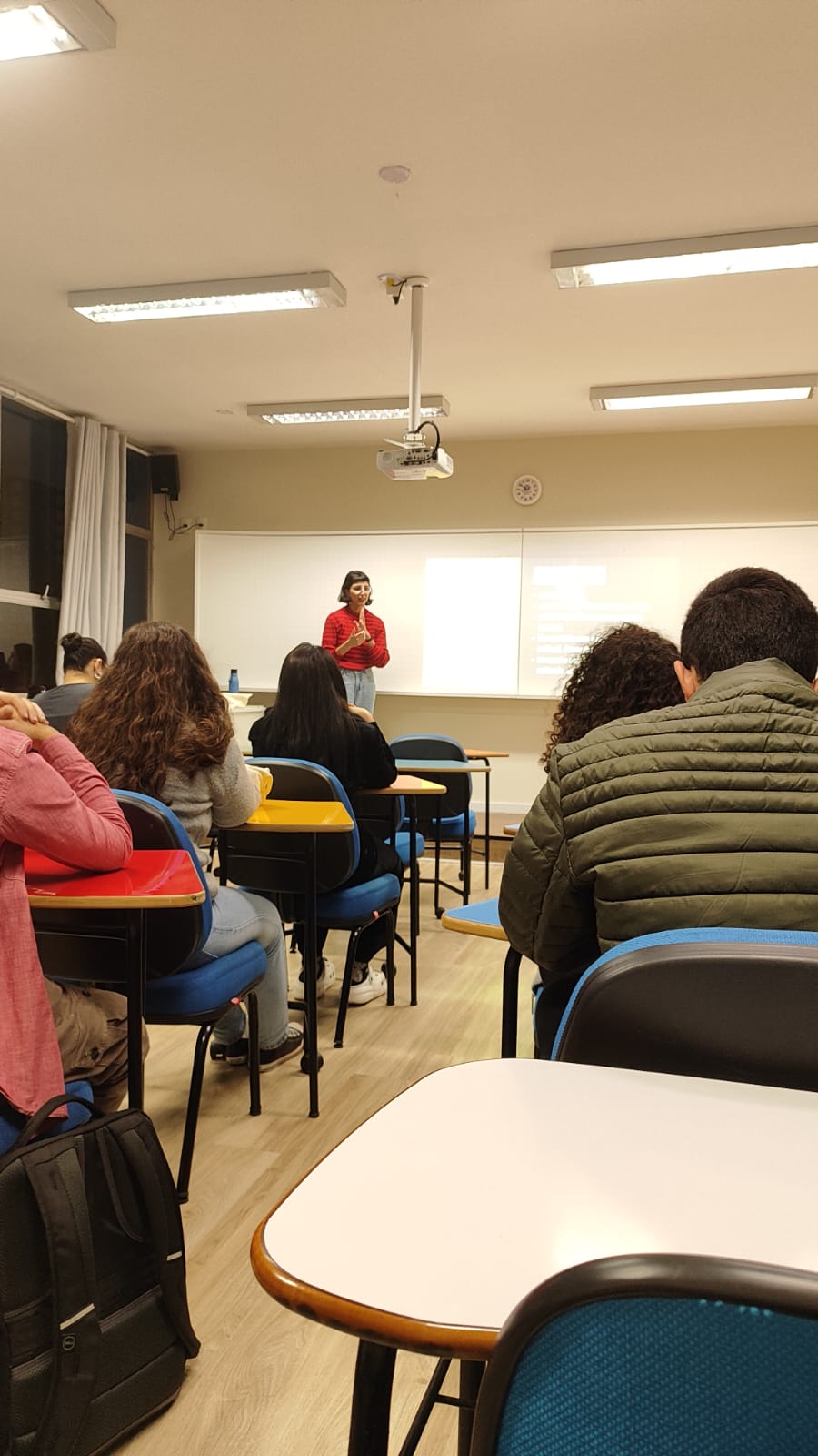 Fotografia de uma sala de aula com estudantes sentados e voltados para a frente. Ao centro, uma mulher veste blusa vermelha e realiza uma apresentação sobre jornalismo cultural. Atrás dela, um slide projetado em uma tela exibe tópicos relacionados ao tema. O ambiente é iluminado e possui quadro branco e projetor no teto.