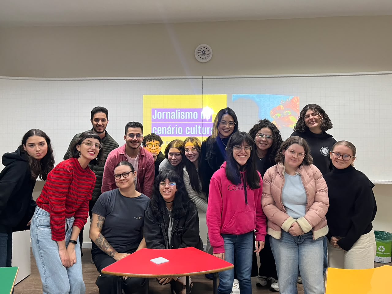Foto em plano conjunto da jornalista Bárbara Siementkowski, de blusa listrada em tons de vermelho e cabelo curto estilo chanel, posando ao lado dos estudantes da oficina de Jornalismo Cultural durante o evento Jornalismo Muda o Mundo. O grupo está reunido em uma sala de aula, sorrindo para a câmera, com uma projeção ao fundo relacionada à atividade.