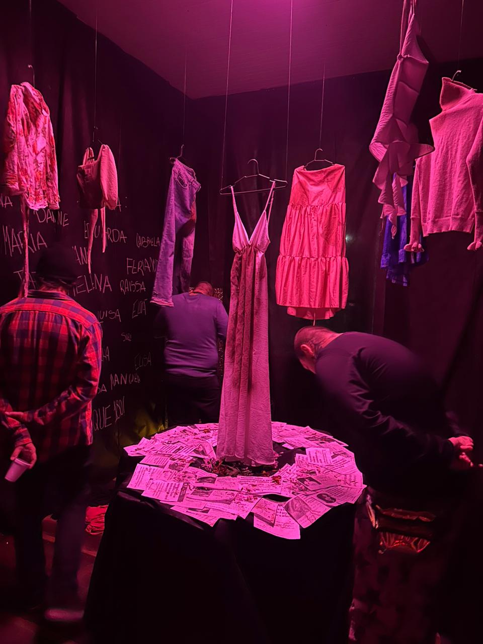 Sala forrada com tecido preto. Em uma das paredes, estão escritos, com giz branco, vários nomes de mulheres. No centro, peças de roupas femininas sujas de sangue penduradas em cabides. Na mesa, no centro da sala, várias notícias impressas com casos de feminicídio e abusos. Sobre os papeis, pétalas de flores espalhadas.