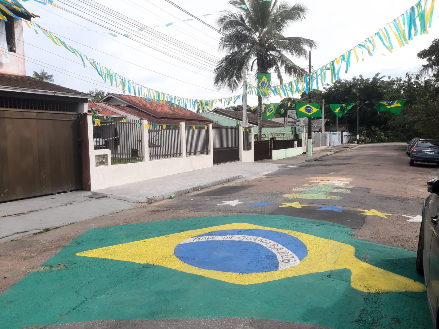 Pintura da bandeira do Brasil realizada no asfalto de uma rua residencial decorada com bandeirolas verde-amarelas 

