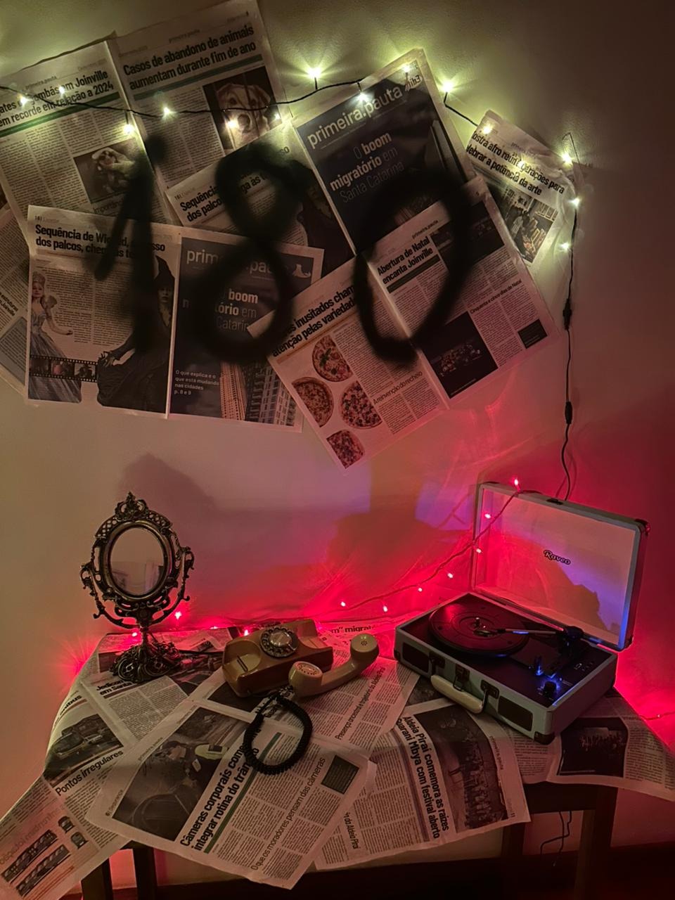 Mesa forrada com jornais. Em cima dela, um espelho antigo, um telefone fora do gancho e uma vitrola que reproduz áudios de mulheres em situação de denúncia. Na parede, folhas de jornais coladas e o número 180 escrito em tamanho grande.