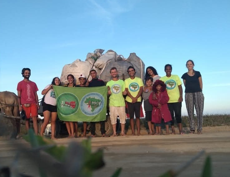 Grupo de voluntários posa para fotografia ao lado de uma carretinha carregada com resíduos recolhidos em mutirão ambiental. Os participantes seguram uma bandeira da associação, enquanto a imagem é enquadrada por vegetação desfocada em primeiro plano.